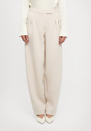 Vrouw in hooggekwaliteit, geplooide beige wijde broek met een witte lange mouw top en witte puntige schoenen tegen een witte achtergrond.