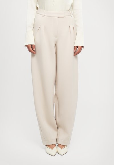 Donna che indossa pantaloni beige a vita alta con pieghe e gamba larga, abbinati a una maglietta bianca a maniche lunghe e scarpe bianche a punta su sfondo bianco.