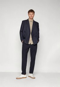 Blazer blu navy a righe sottili doppiopetto indossato sopra una camicia beige con motivo, abbinato a pantaloni neri e sneakers bianche con dettagli color cuoio.