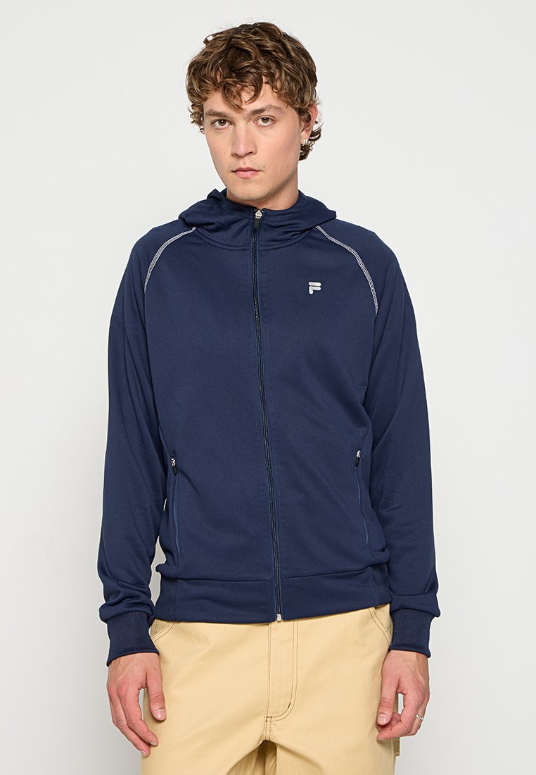 Felpa zip con cappuccio color navy, colletto alto, polsini elastici e due tasche laterali. Presenta cuciture riflettenti e un logo sul lato sinistro del petto.