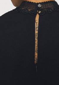 Dos d'une robe noire avec col en dentelle et petits boutons, montrant une fermeture éclair et un tatouage vertical sur le cou du porteur.