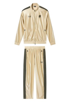 Tuta beige lucida con righe verticali nere sulle maniche e sui pantaloni, giacca con zip frontale, polsini elastici e loghi EA7 su giacca e pantaloni.