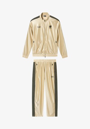 Tuta beige lucida con righe verticali nere sulle maniche e sui pantaloni, giacca con zip frontale, polsini elastici e loghi EA7 su giacca e pantaloni.