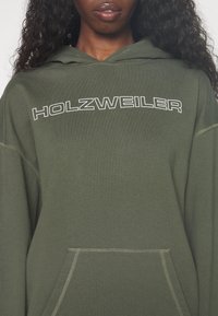 Femme portant un sweat à capuche vert olive avec le mot HOLZWEILER imprimé en blanc sur la poitrine.