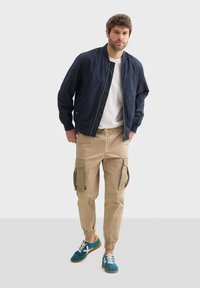 Námorní plášť s modrými akcenty, bílým tričkem, béžovými cargo kalhotami s bočními kapsami a modrými teniskami s bílými tkaničkami a hnědými podrážkami.