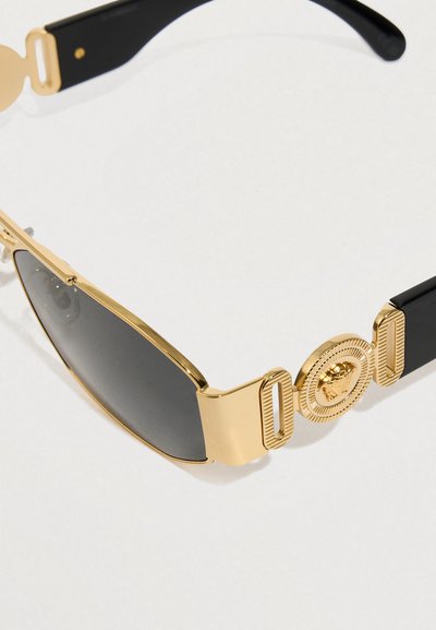 Čierne slnečné okuliare so zlatým kovovým rámom, s okrúhlym emblémom a textúrovanými detailmi na stranách. Obsahujú tmavo tónované šošovky.