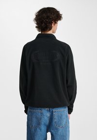 Sort fleece-pullover med krave, der har et præget logo på ryggen, parret med blå denimjeans. Blød tekstur og afslappet pasform.