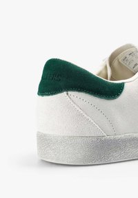Zapatillas de ante blancas con un talón de ante verde y suela gris texturizada. Presenta un sutil logo grabado en el talón. Diseño sencillo.