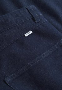 Dunkelblaue Denimstoff mit einer Tasche, die ein weißes Etikett mit der Aufschrift "GABBA" trägt. Glatte Textur und saubere Nähte sind sichtbar.