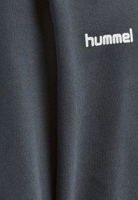 Donkergrijze stof met een gladde textuur, met het witte merklogo "hummel" afgedrukt nabij de bovenkant.