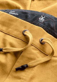 Tissu en velours côtelé jaune avec des cordons tressés marron et des œillets en métal. La doublure intérieure présente un motif noir avec des accents rouges.