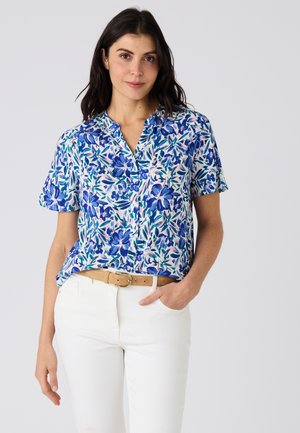 Mujer de cabello oscuro vistiendo una blusa de manga corta con flores azules y blancas, cinturón beige y pantalones blancos, de pie con la mano en el bolsillo.