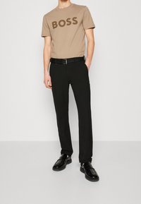 Beige bomullst-shirt med mörkbrun "BOSS"-logotyp, kombinerad med svarta skräddarsydda byxor och svarta läderskor. Enkel, modern design.