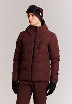 Man draagt een donkerrode winterjas, bijpassende broek, zwarte handschoenen, een bordeauxrode muts en zwarte skibril op het hoofd, kijkt naar links.