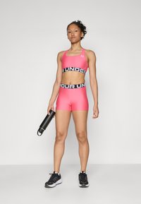 Rosa sport-bh och short-set med svart elastisk midja med texten "UNDER". Modellen håller en svart vattenflaska. Svarta sneakers.