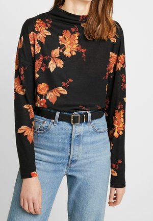 Schwarzes langärmeliges Oberteil mit floralem Muster in Orange und Gelb, kombiniert mit hoch geschnittenen blauen Jeans und einem schwarzen Gürtel mit einer goldenen Schnalle.