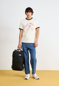 Tricou crem cu margini negre, având imprimeu "Crazy Cougars", blugi albaștri, sneakers albi și un rucsac negru North Face.