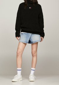 Person som bär en svart polotröja, ljusblå asymmetrisk jeanskjol, vita sneakers och vita strumpor med marinblå ränder.