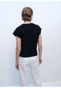 Black cotton t-shirt with short sleeves and a fitted waist design, paired with white pants, shown from the back against a neutral background.