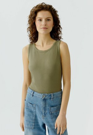 Frau mit lockigem braunem Haar, die ein olivgrünes ärmelloses, geripptes Oberteil und hellblaue, hoch taillierte Jeans mit großen Vorderentaschen trägt.