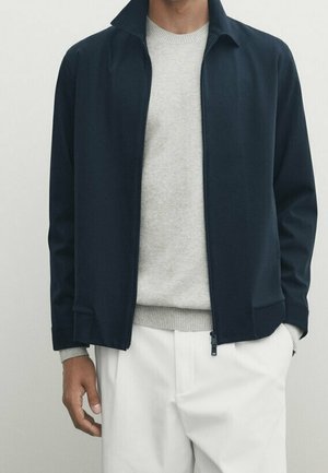 Mand iført en navyblå lynlåstrøje over en lysegrå sweater og hvide bukser, stående mod en ensfarvet lys baggrund.