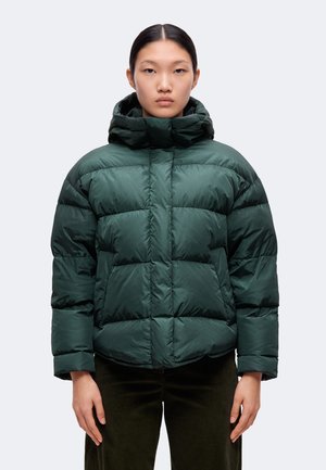 Femme aux cheveux noirs portant une veste matelassée vert foncé avec un col montant et des manches longues, debout devant un fond clair uni.
