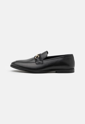 Mocassins en cuir noir avec une texture lisse, un bout arrondi et une boucle décorative dorée sur le dessus. Design minimaliste.