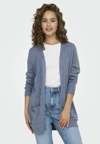 Světle modrý cardigan vyrobený z měkkého materiálu, s dlouhými rukávy, dvěma předními kapsami a žebrovanými manžetami. Nosit na bílém tričku a džínách.