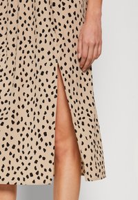 Knee-length, béžové šaty s černým tečkovaným vzorem, se bočním rozparkem. Materiál vypadá lehký a má hladkou texturu.