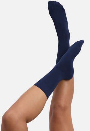 Benen omhoog, gedragen met effen marineblauwe sokken tot halverwege de kuit tegen een effen lichte achtergrond.