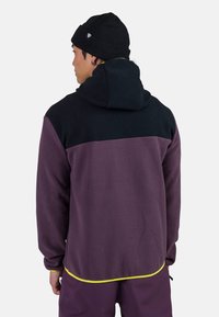 Il pullover in pile presenta un design a blocchi di colore con sezioni nere e prugna, un cappuccio e un bordo giallo lungo l'orlo.