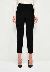 Black tailored trousers with a high waist, featuring front pleats, side pockets, and a slim-fit design, paired with black pointed heels.