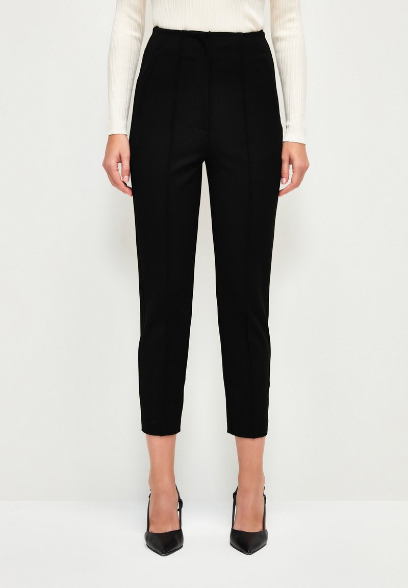 Black tailored trousers with a high waist, featuring front pleats, side pockets, and a slim-fit design, paired with black pointed heels.