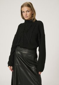 Schwarzer Strickpullover mit hohem Kragen und strukturierten Details, kombiniert mit einem schwarzen Leder-Midikleid mit glatter Oberfläche.