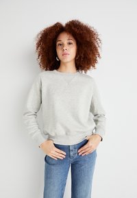 Sweat-shirt gris avec un col rond et des poignets côtelés. Présente une texture subtile, cintré à la taille, porté avec un jean bleu taille haute.