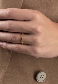Bague en or avec un design texturé et perlé. Portée sur une main contre un fond en tissu brun avec un détail de bouton visible.
