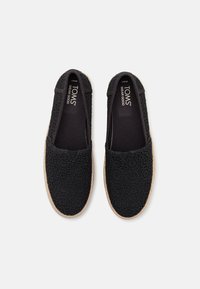 Zapatos negros tipo slip-on con un patrón de encaje floral, acentos elásticos en los laterales y una suela texturizada estilo espadrille. Marca visible en el interior.