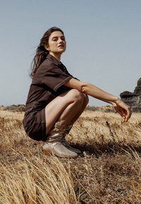 Botas de tobillo plateadas metálicas con un diseño arrugado, combinadas con un traje de color marrón oscuro y de manga corta. El fondo presenta hierba seca y rocas.