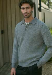 Suéter gris de punto con cuello tipo polo, tres botones y textura acanalada; llevado sobre una camisa blanca, combinado con pantalones oscuros.