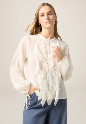 Blusa bianca con frontale arricciato, scollatura rotonda e chiusura con bottoni. Maniche lunghe, leggermente trasparenti, con polsini arricciati; abbinata a pantaloni blu.