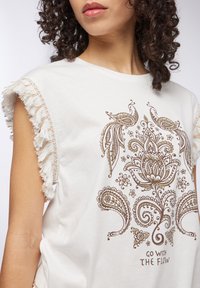 T-shirt bianco leggero con un design floreale e paisley marrone, con bordo frangiato lungo le maniche e una vestibilità ampia.