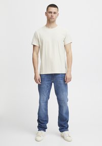 Bež majica od pamuka s okruglim izrezom, u kombinaciji s izblijedjelim plavim trapericama. Model u off-white tenisicama stoji protiv jednostavne pozadine.