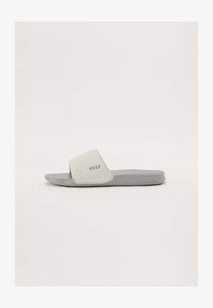 Sandal cinza com uma tira texturizada branca. A tira apresenta perfurações e está marcada com o nome da marca "REEF". Sola plana, design minimalista.