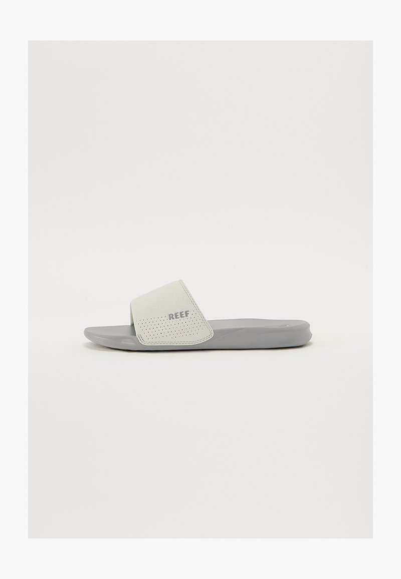 Graue Rutschsandale mit einem strukturierten weißen Riemen. Der Riemen ist durchlöchert und trägt den Markennamen "REEF". Flache Sohle, minimalistisches Design.