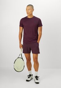 Homme en t-shirt et short de sport bordeaux tenant une raquette de tennis, portant des chaussettes blanches et des chaussures de tennis blanches, se tenant devant un fond uni.