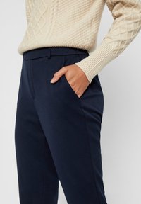 Marina skräddarsydda byxor med en slät textur, utrustade med sidofickor och en rak framdesign, kompletterade av en krämfärgad kabelstickad tröja.