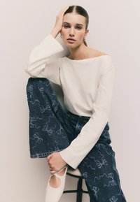 Weißes Langarmoberteil, dunkelblaue weite Jeans mit Schleifenmuster und weiße Schuhe mit Ausschnitt-Details. Model sitzt auf einem Hocker.