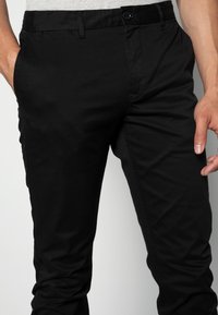 Pantaloni in cotone neri con una vestibilità slim, dotati di chiusura con bottone e tasche laterali. Il tessuto ha una consistenza liscia e una leggera lucentezza.