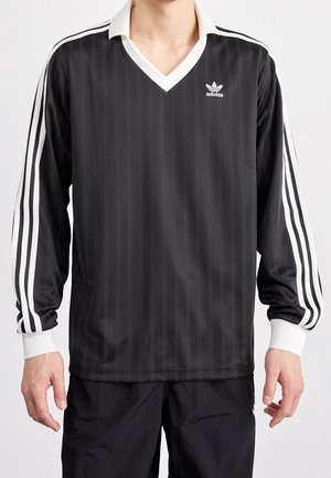 Mann trägt ein schwarzes Adidas Langarm-Sportshirt mit weißen Streifen an den Ärmeln und weißem Kragen vor einfachem Hintergrund.