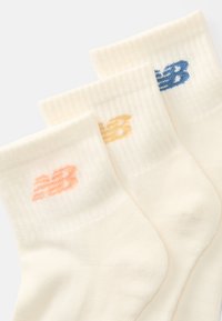 Trois paires de chaussettes blanches à côtes, avec le logo "NB" en orange, jaune et bleu sur les bords, disposées en superposition sur une surface blanche.